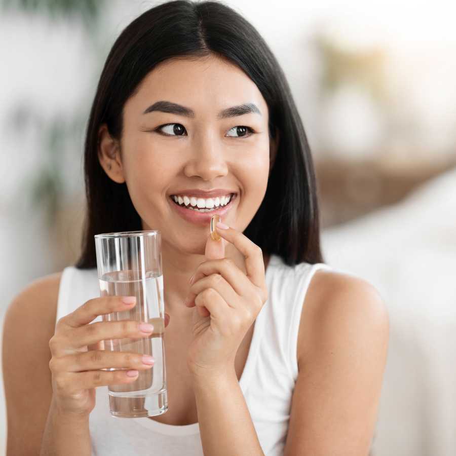 smiling woman holding a glass of water in one hand and a supplementary capsule in the other